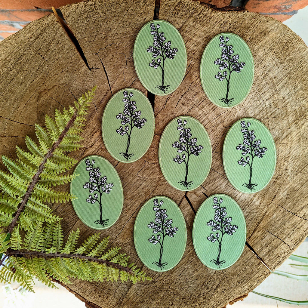 Eight oval iron-on patches from ilikesara | art + goods, each with a botanical illustration, are arranged on a tree stump slice. An Iron-on patch | maidenhair fern is displayed to the left of the other patches.