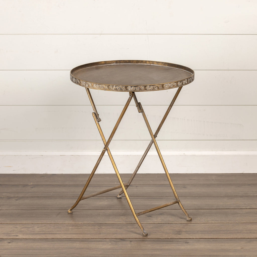 The Ragon House ROUND GOLD FOLDING TRAY TABLE features a distressed gold finish and foldable crossed legs, making it ideal as a lightweight table or plant stand on a wooden floor against a white shiplap wall.