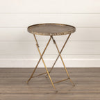 The Ragon House ROUND GOLD FOLDING TRAY TABLE features a distressed gold finish and foldable crossed legs, making it ideal as a lightweight table or plant stand on a wooden floor against a white shiplap wall.