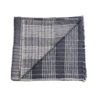 A folded Silica Rose Throw by Variously, in shades of salmon with a gray stripes pattern, is shown with one corner flipped over to reveal the reverse side. This handwoven piece features a linear striped design and frayed edges.