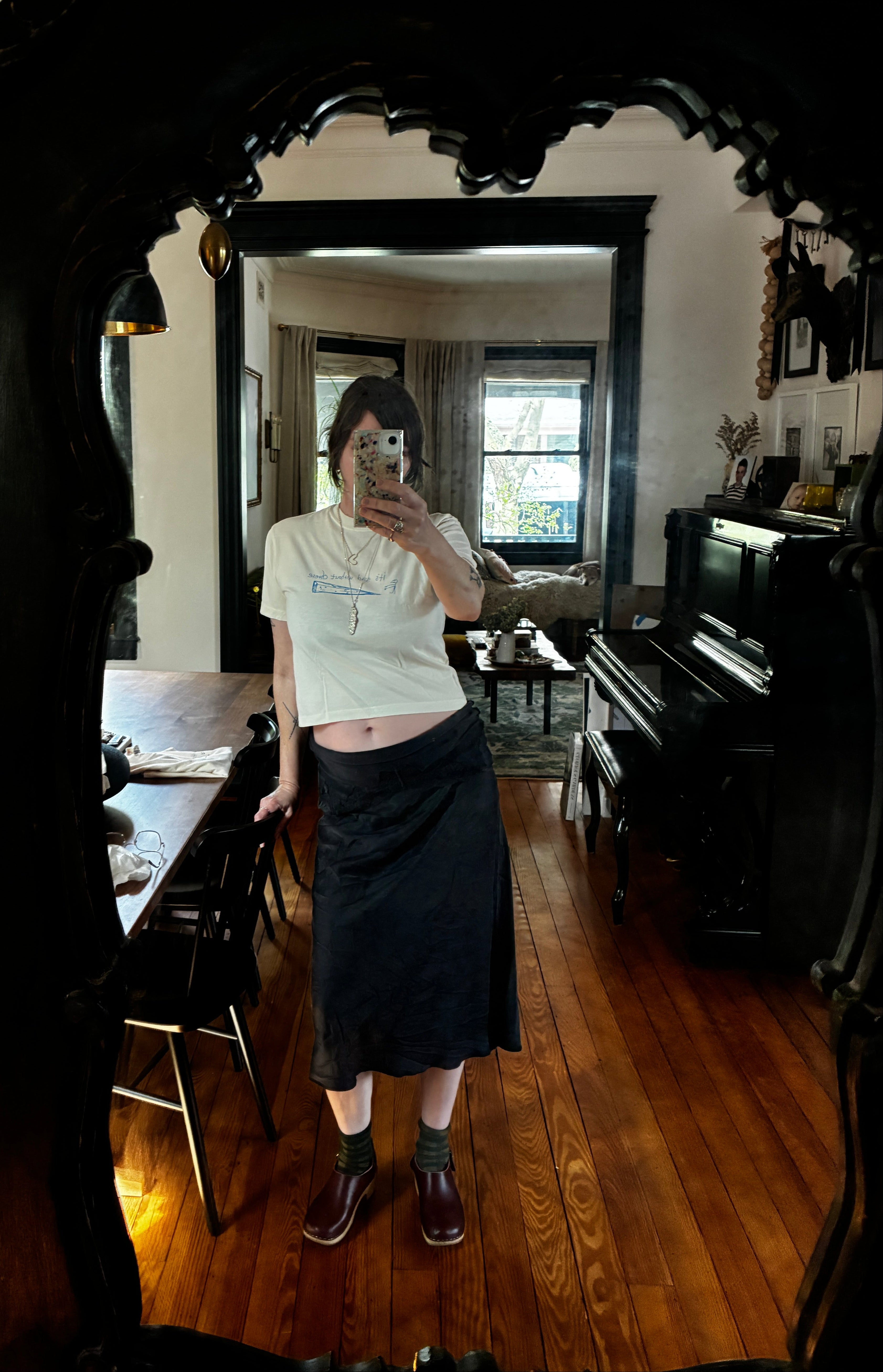 Person taking a mirror selfie in a room with wooden floors, wearing the limited edition Thyne Goods "Cheese" 100% cotton white t-shirt, dark skirt, socks, and brown shoes. The room has a piano, table, chairs, and a window showing trees outside.