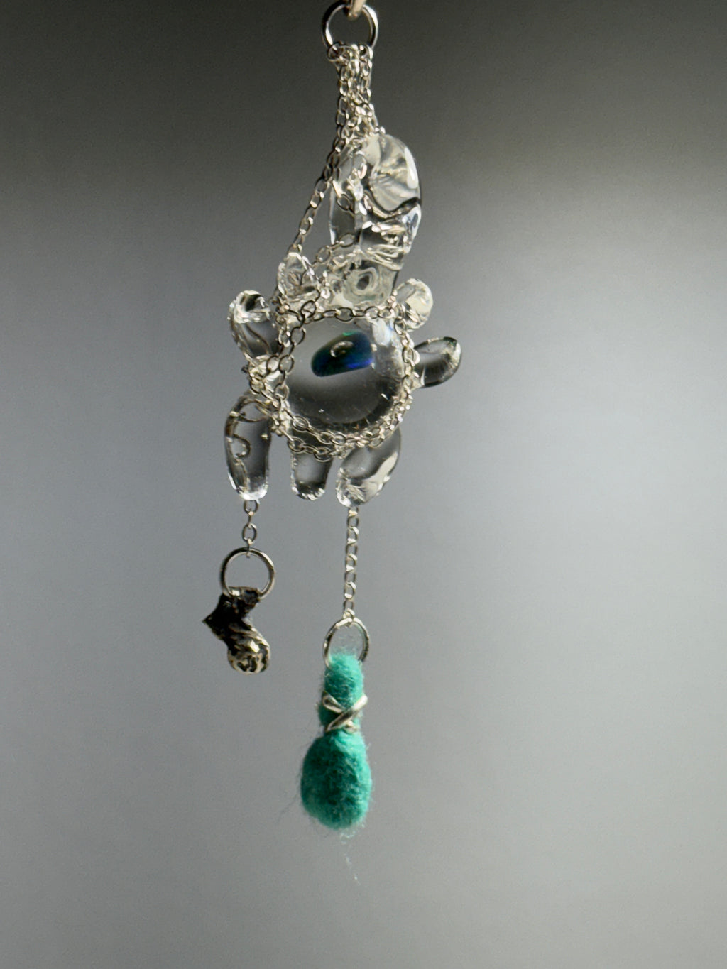 Catch • Ash Espinoza x Thyne Studio by Thyne Goods is a delicate pendant with silver wire, a clear bead, blue-green eye center, and teal felted bead, highlighting recycled sterling silver jewelry against a plain gray background.