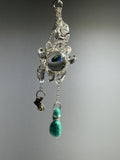 Catch • Ash Espinoza x Thyne Studio by Thyne Goods is a delicate pendant with silver wire, a clear bead, blue-green eye center, and teal felted bead, highlighting recycled sterling silver jewelry against a plain gray background.