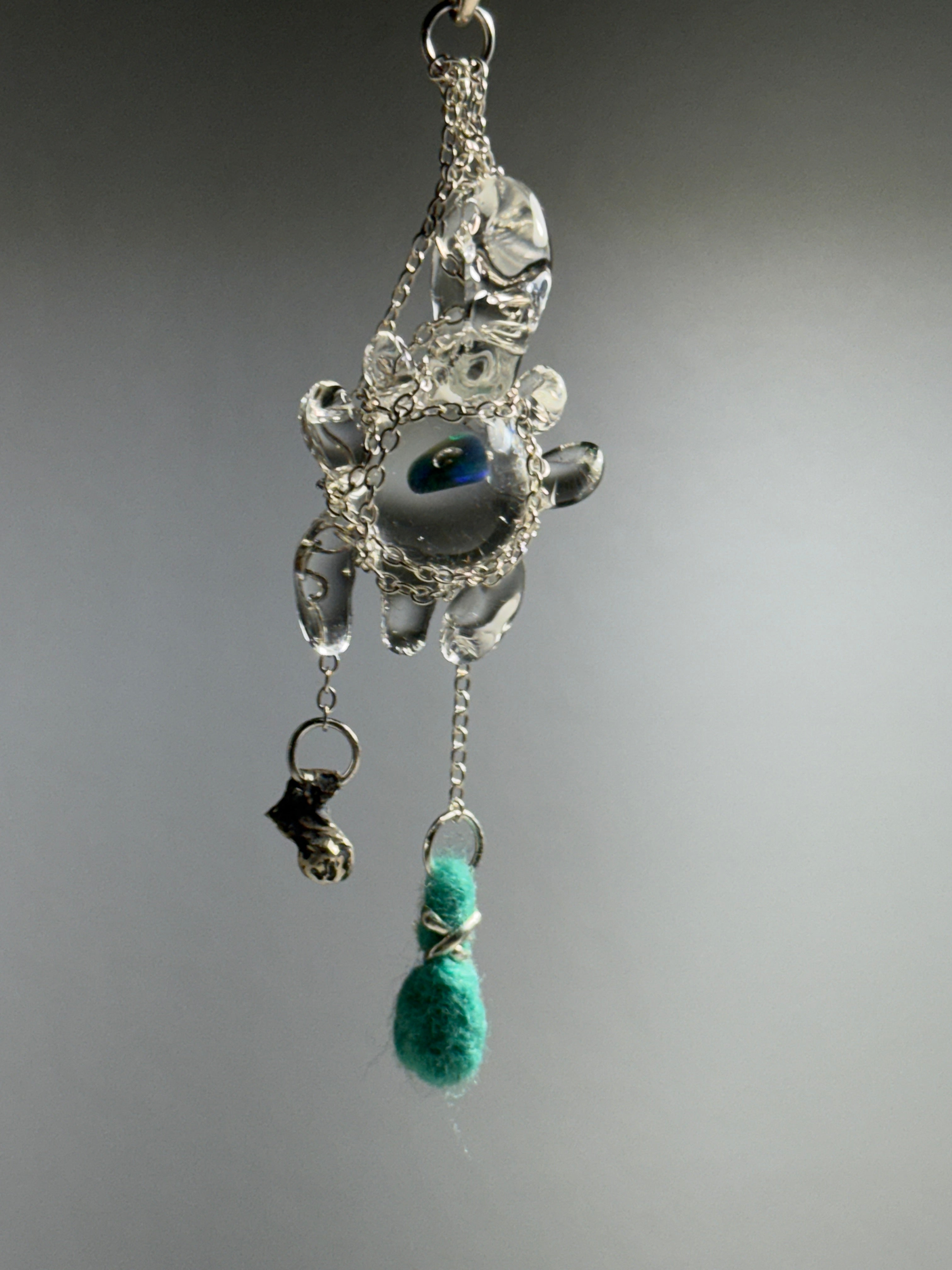 Catch • Ash Espinoza x Thyne Studio by Thyne Goods is a delicate pendant with silver wire, a clear bead, blue-green eye center, and teal felted bead, highlighting recycled sterling silver jewelry against a plain gray background.