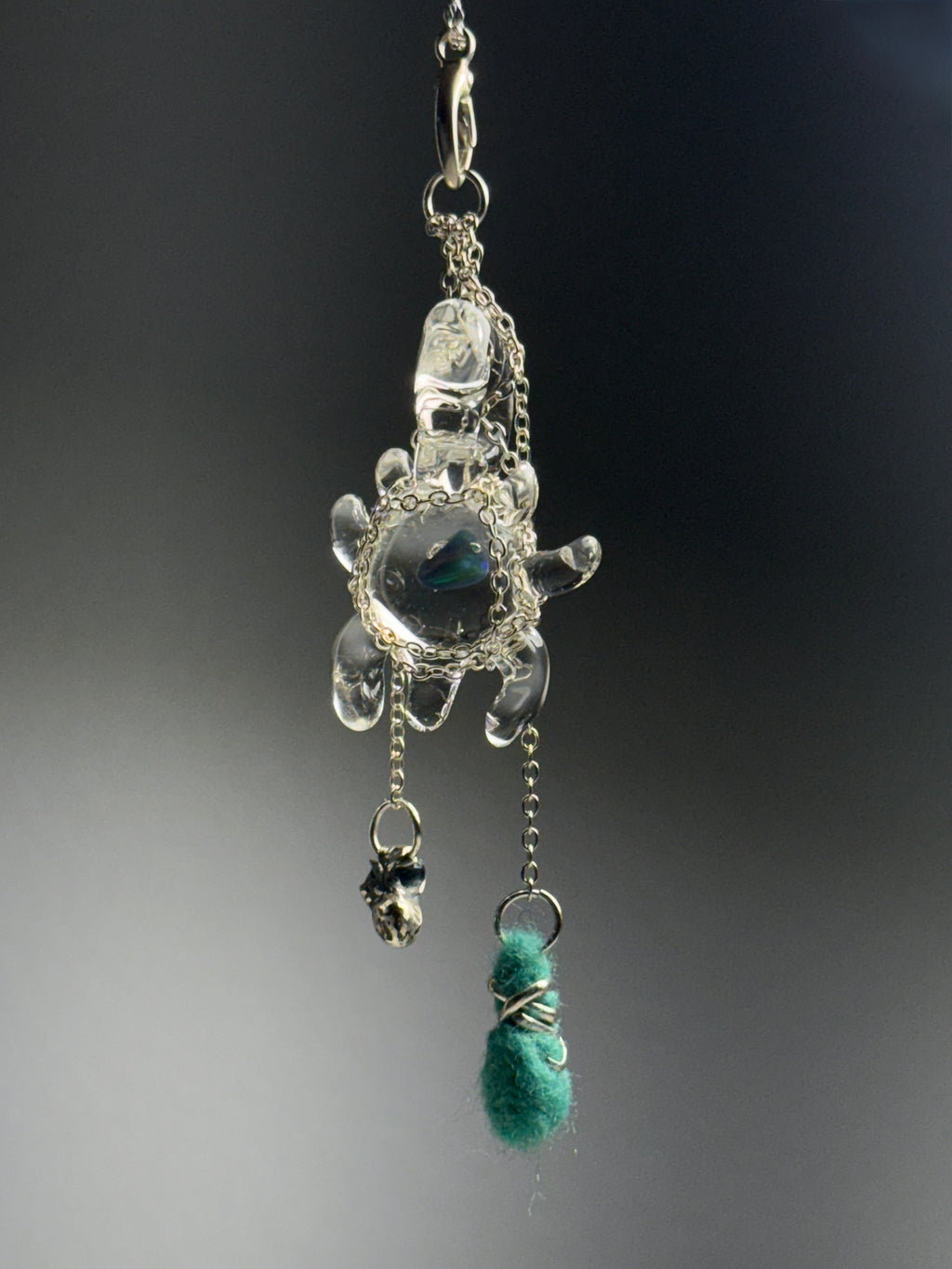 Catch • Ash Espinoza x Thyne Studio by Thyne Goods: A turtle-shaped recycled silver pendant on a chain, adorned with a small silver skull charm and a green fuzzy wire-wrapped bead, set against a dark, blurred background.