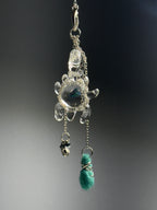 Catch • Ash Espinoza x Thyne Studio by Thyne Goods: A turtle-shaped recycled silver pendant on a chain, adorned with a small silver skull charm and a green fuzzy wire-wrapped bead, set against a dark, blurred background.