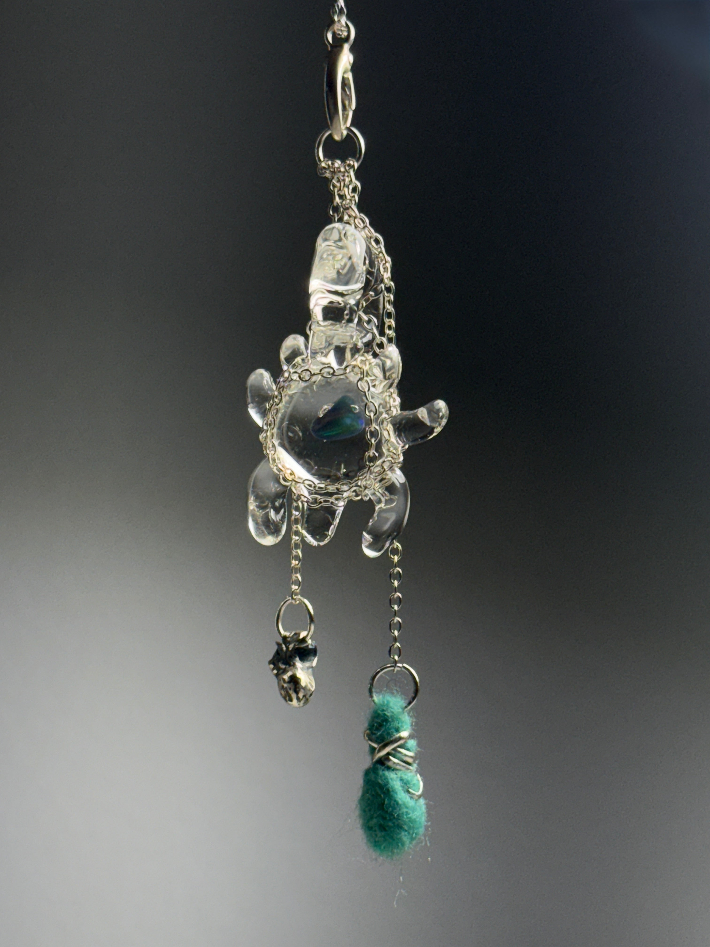 Catch • Ash Espinoza x Thyne Studio by Thyne Goods: A turtle-shaped recycled silver pendant on a chain, adorned with a small silver skull charm and a green fuzzy wire-wrapped bead, set against a dark, blurred background.