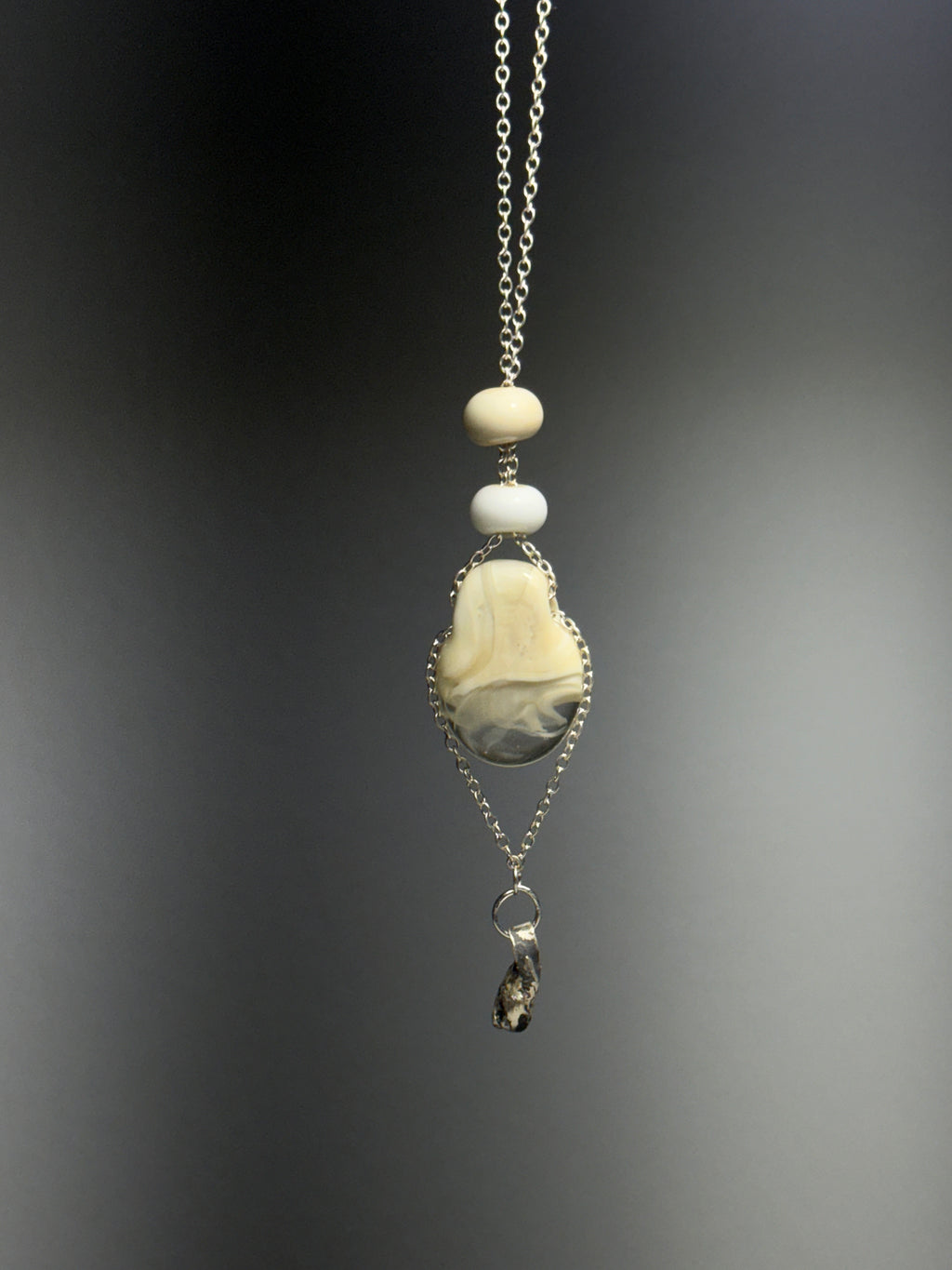 The Dove necklace by Thyne Goods (Ash Espinoza x Thyne Studio) features a silver chain with cream and white beads, a marbled cream-gray stone in recycled sterling silver, and a small dark pendant. Softly blurred background.