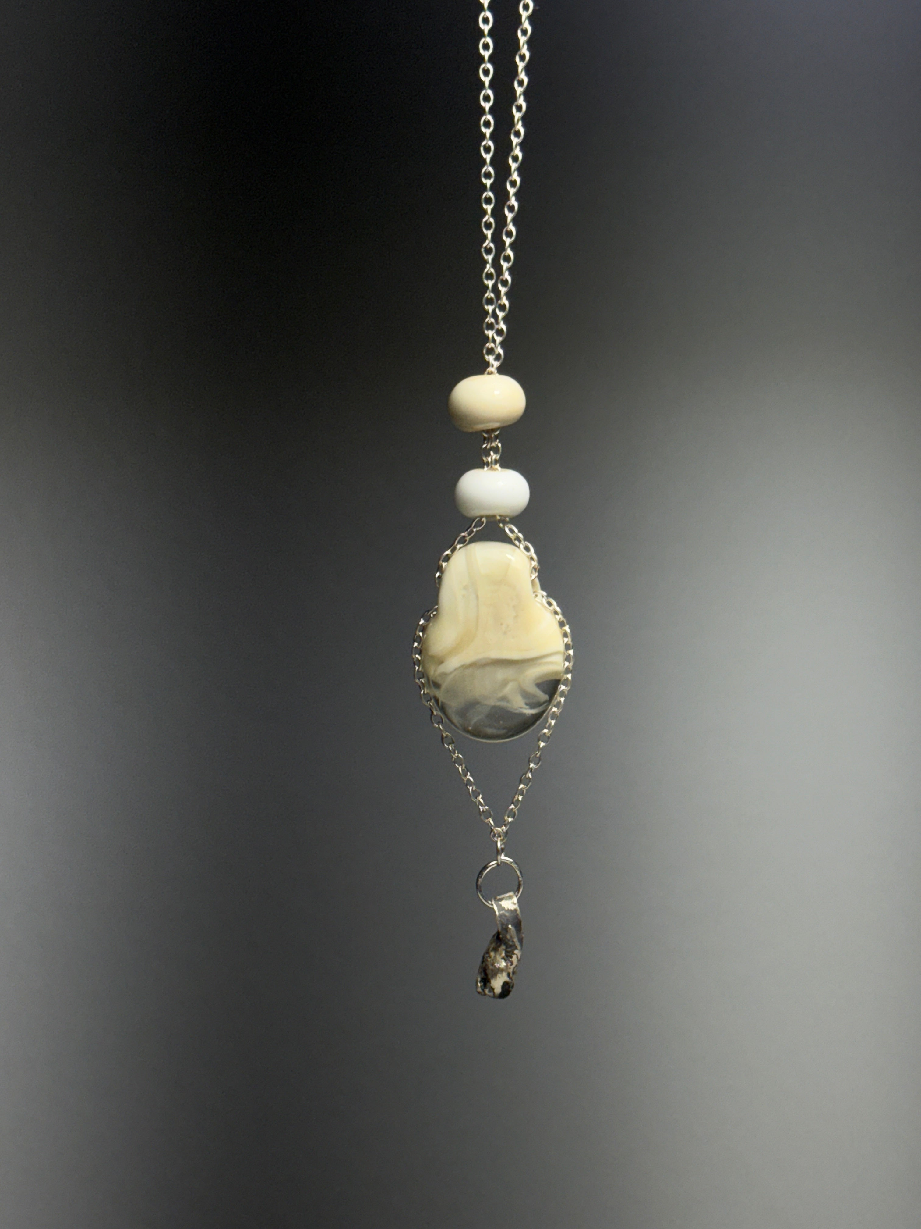 The Dove necklace by Thyne Goods (Ash Espinoza x Thyne Studio) features a silver chain with cream and white beads, a marbled cream-gray stone in recycled sterling silver, and a small dark pendant. Softly blurred background.