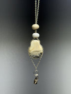 Dove by Ash Espinoza x Thyne Studio for Thyne Goods: a pendant necklace with two round white glass beads, a polished oval cream and gray stone, an irregular dark charm, all on recycled sterling silver chains over a gray gradient backdrop.