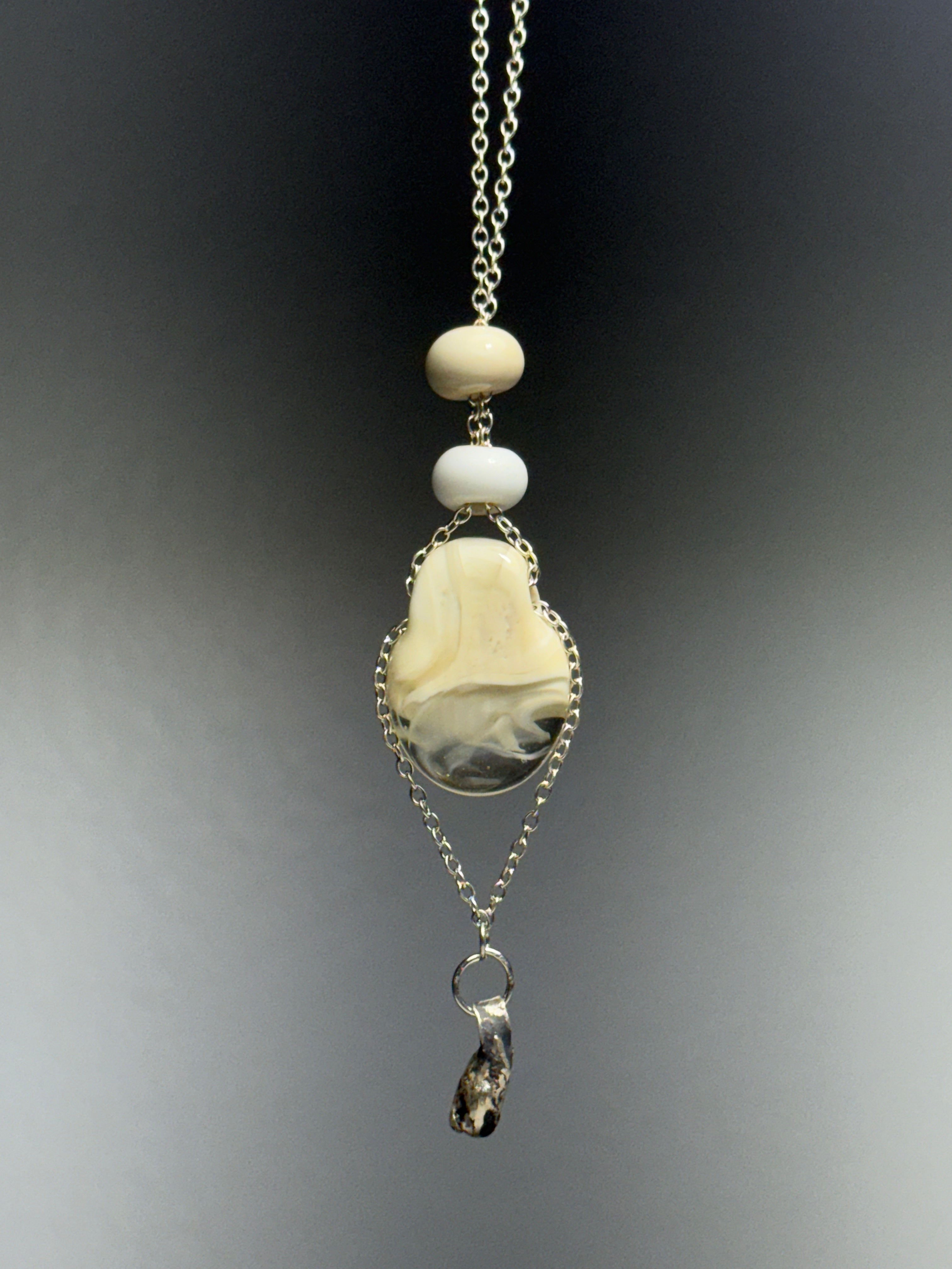 Dove by Ash Espinoza x Thyne Studio for Thyne Goods: a pendant necklace with two round white glass beads, a polished oval cream and gray stone, an irregular dark charm, all on recycled sterling silver chains over a gray gradient backdrop.