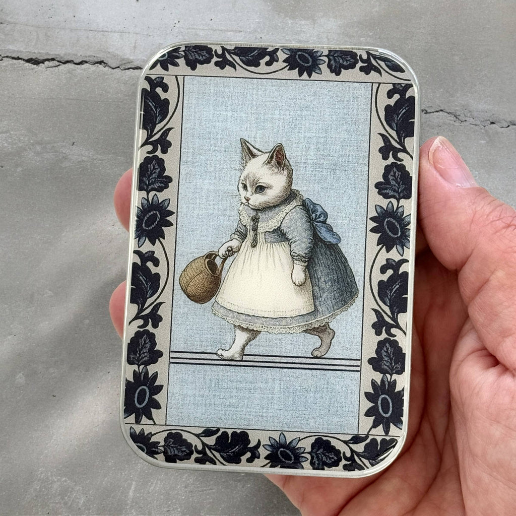A hand holds the Firefly Notes Kitty notions tin: Small, featuring a white business cat in a dress and apron carrying a basket, surrounded by dark floral borders. The rectangular tin clicks shut securely.