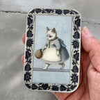 A hand holds the Firefly Notes Kitty notions tin: Small, featuring a white business cat in a dress and apron carrying a basket, surrounded by dark floral borders. The rectangular tin clicks shut securely.