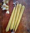 A pair of 7 inch hand-dipped pure beeswax tapers from Yarrow Bone Farmstead rest on a wooden surface beside dried white flowers, with one candle's wick extended and linked to another.