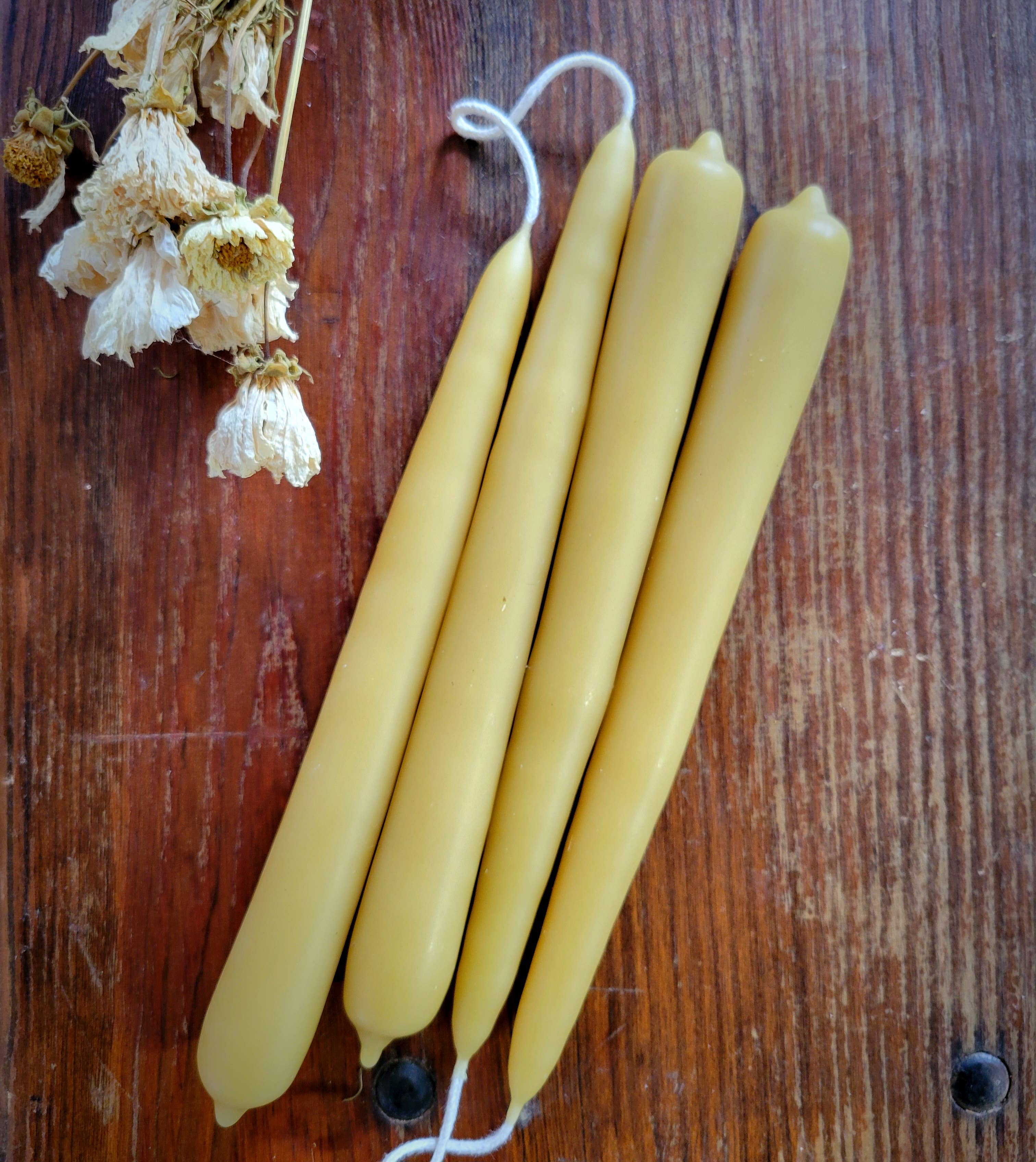 A pair of 7 inch hand-dipped pure beeswax tapers from Yarrow Bone Farmstead rest on a wooden surface beside dried white flowers, with one candle's wick extended and linked to another.