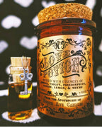 A North Ave Candles "The Apothecary / Love Potion / Valentine Candle" in a vintage-style glass jar with cork lid sits next to a small vial topped with a key-shaped charm, set against a patterned background, evoking an air of mystery.