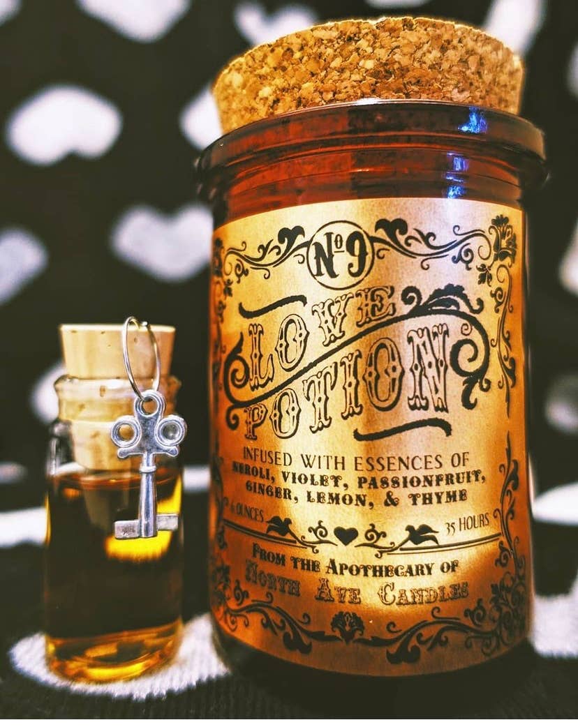 A North Ave Candles "The Apothecary / Love Potion / Valentine Candle" in a vintage-style glass jar with cork lid sits next to a small vial topped with a key-shaped charm, set against a patterned background, evoking an air of mystery.