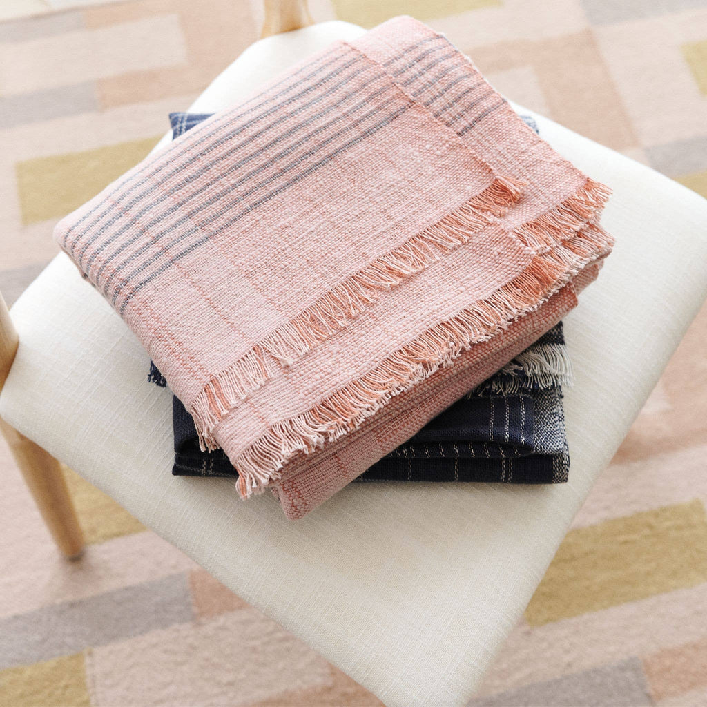 The Silica Rose Throw in shades of salmon with gray stripes by Variously is folded atop other dark-hued fabrics, all stacked neatly on a white cushioned seat. Fringed edges add artisanal detail, while a softly patterned carpet in the background complements this handcrafted textile.
