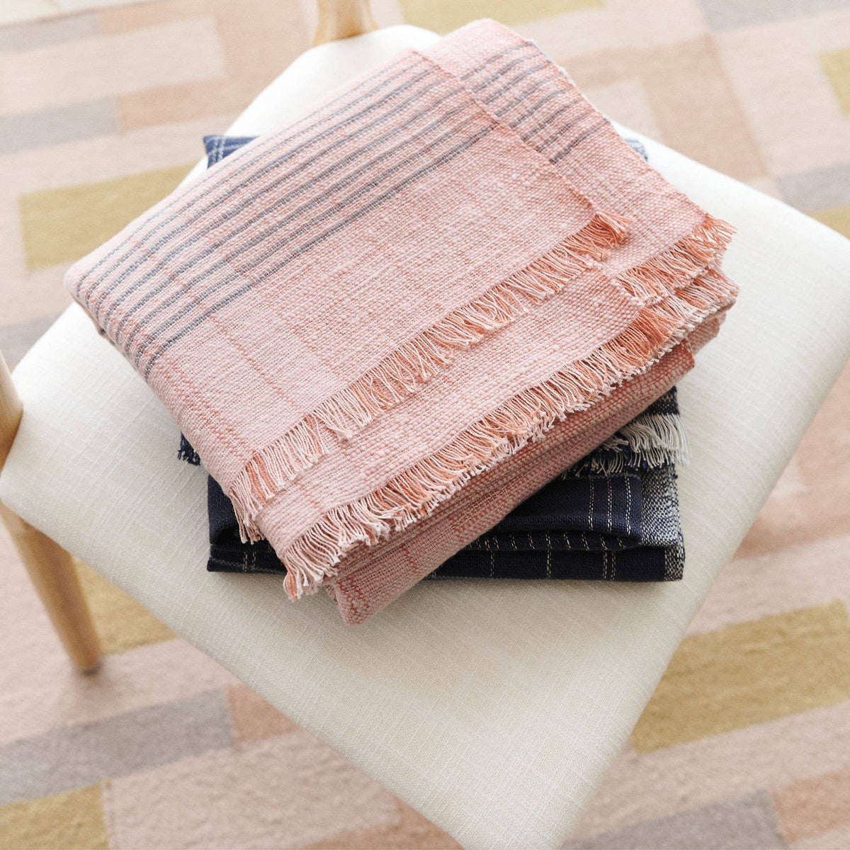 The Silica Rose Throw in shades of salmon with gray stripes by Variously is folded atop other dark-hued fabrics, all stacked neatly on a white cushioned seat. Fringed edges add artisanal detail, while a softly patterned carpet in the background complements this handcrafted textile.