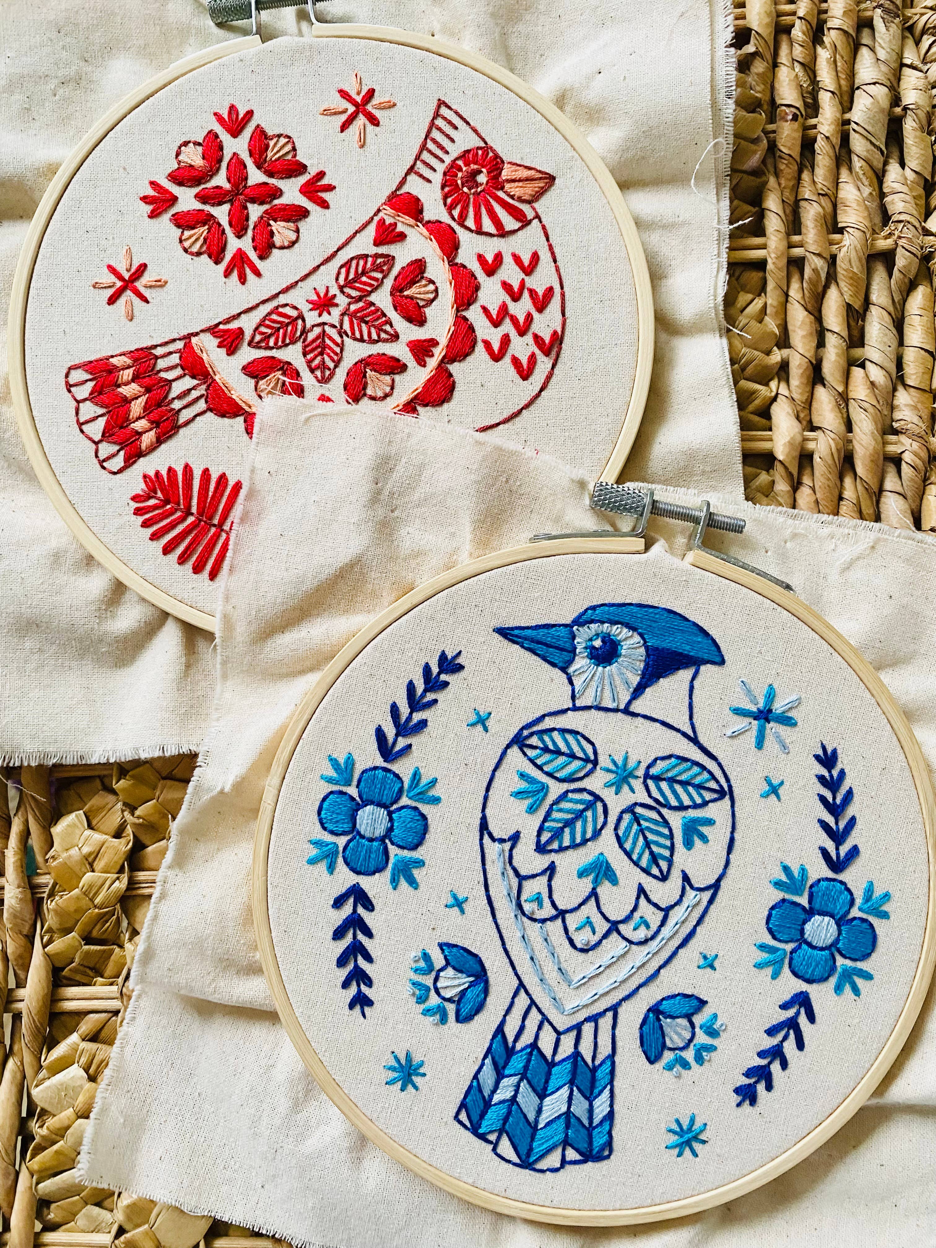 Two fabric pieces in hoops display vibrant designs: Hook, Line & Tinker’s Blue Jay Complete Embroidery Kit—one with a red bird among flowers, the other a blue jay with floral patterns—both ideal for beginner embroiderers.