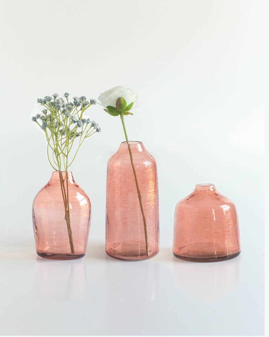 Three Bella Handblown Hammered Glass Bud Vase: Blush by Creative Women are lined up, with white blooms in the left vase, a single flower in the middle, and the right vase empty against a plain white background.