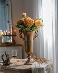 A Gold Trophy Vase with Handles by Vagabond Vintage holds yellow and orange peonies on a wooden table by a window with sheer curtains. Nearby are a wine glass, crown, book, dried flowers, and a lit candelabra in the background.