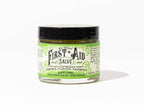 A small glass jar of Roots And Leaves First Aid Salve with a black lid and cream-green label, made with comfrey, plantain, calendula, manuka honey, black cumin seed oil, and hemp seed oil—petroleum free.