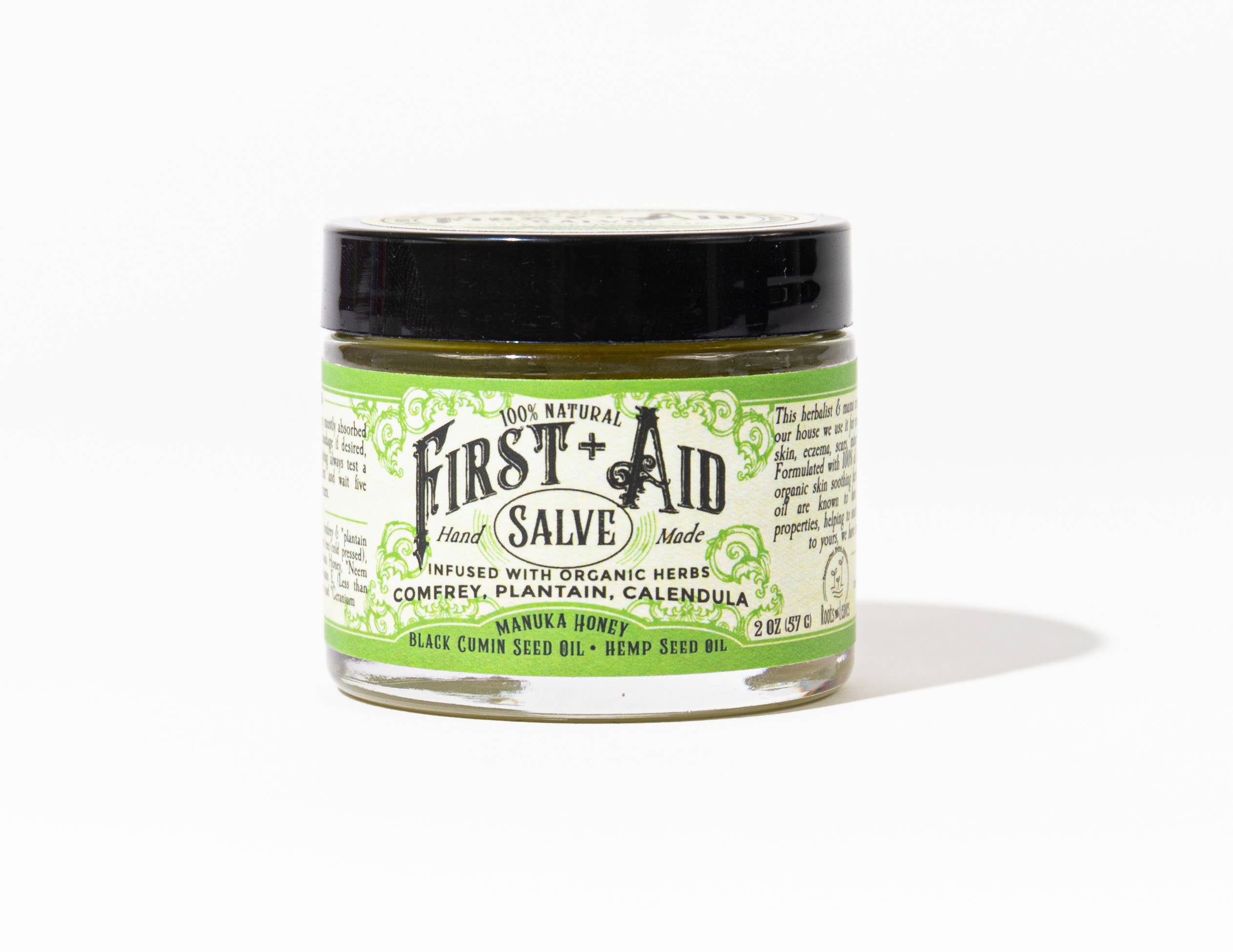 A small glass jar of Roots And Leaves First Aid Salve with a black lid and cream-green label, made with comfrey, plantain, calendula, manuka honey, black cumin seed oil, and hemp seed oil—petroleum free.