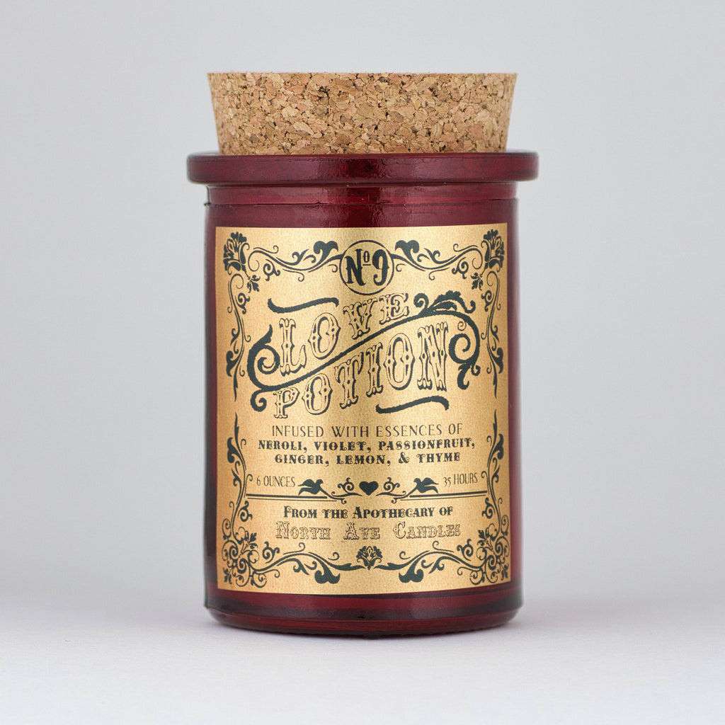 The Apothecary / Love Potion / Valentine Candle by North Ave Candles is a red glass jar candle with a cork lid. Vintage label features neroli, violet, passionfruit, ginger, lemon, and thyme fragrance and a 35-hour burn time.