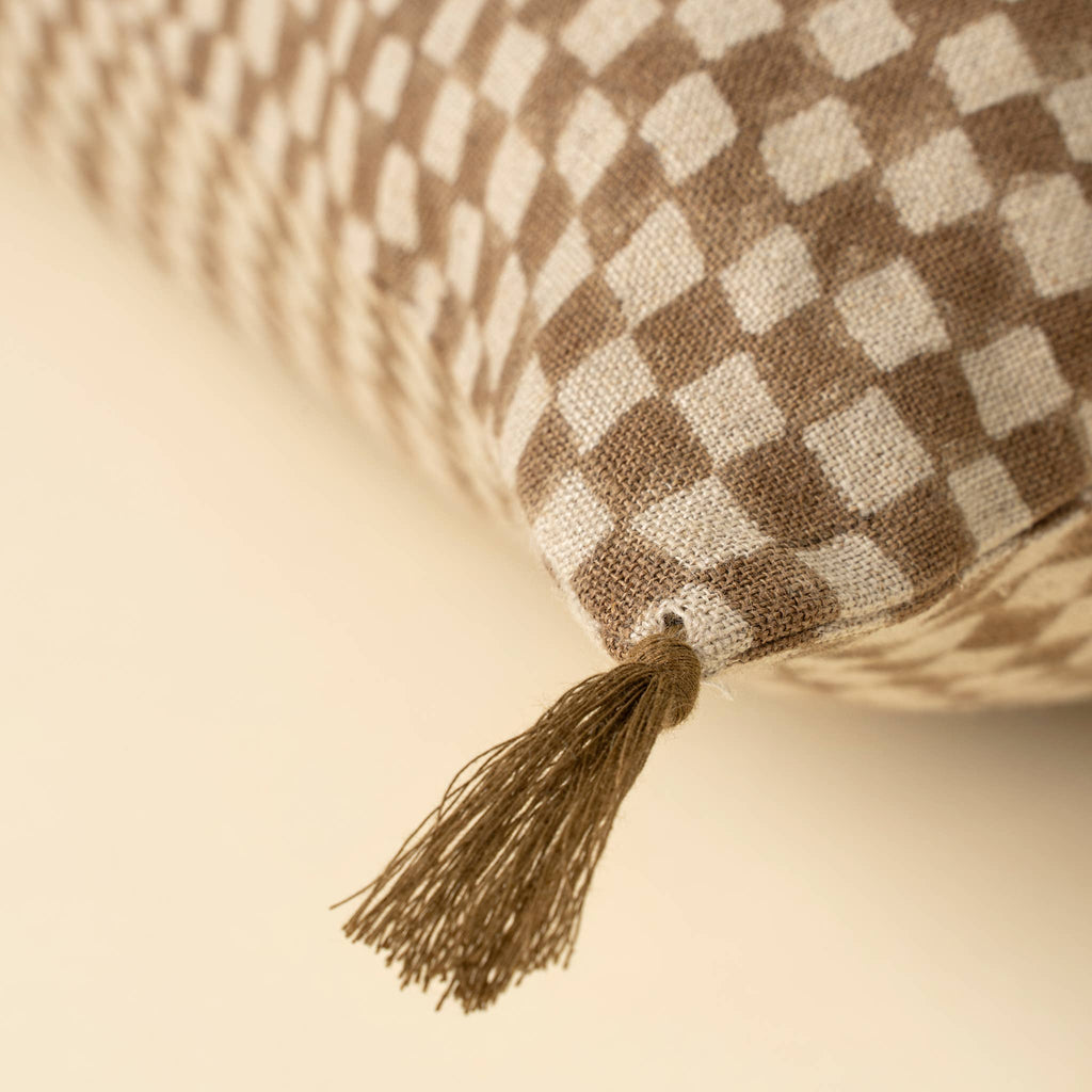 Close-up of the Indaba Trading 16x24 Block Print Check Linen Pillow in brown, featuring a beige and brown checkered design with a fabric tassel at the corner, resting on a light surface. Cover with filler included.