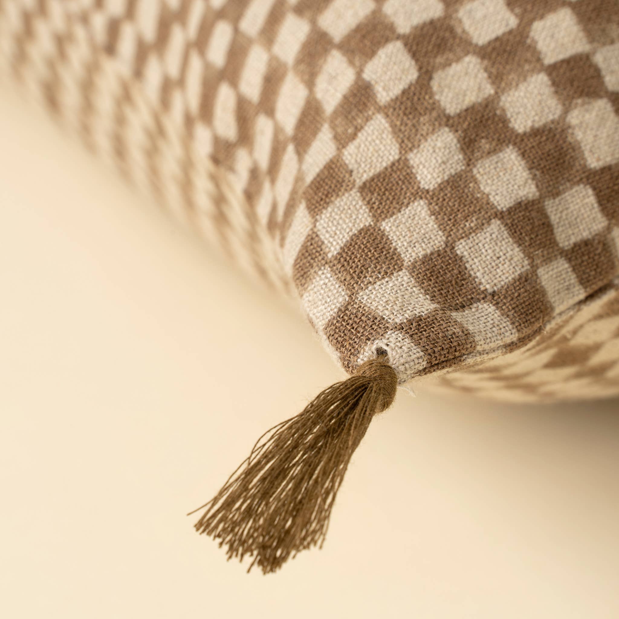 Close-up of the Indaba Trading 16x24 Block Print Check Linen Pillow in brown, featuring a beige and brown checkered design with a fabric tassel at the corner, resting on a light surface. Cover with filler included.