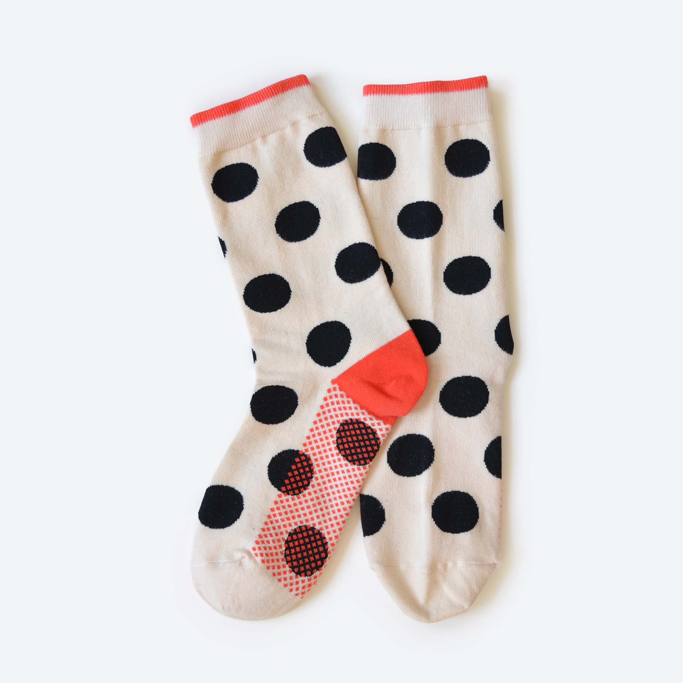 The Valley Sock by Hooray Sock Co. is a unisex cream crew sock with large black polkadots, red heel, toe, and cuff. Made from cotton spandex, it features a red non-slip grip sole for added safety.