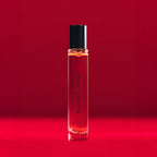 A clear glass SCANDALWOOD ROLLERBALL: 10ML by Heretic Parfum, with a black cap, stands upright against a vibrant red background, evoking hints of sandalwood and musk.