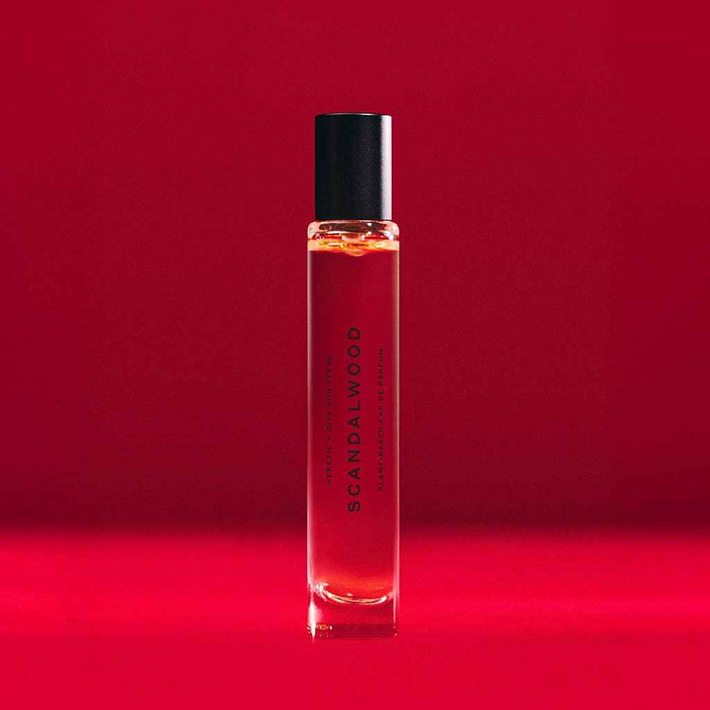 A clear glass SCANDALWOOD ROLLERBALL: 10ML by Heretic Parfum, with a black cap, stands upright against a vibrant red background, evoking hints of sandalwood and musk.