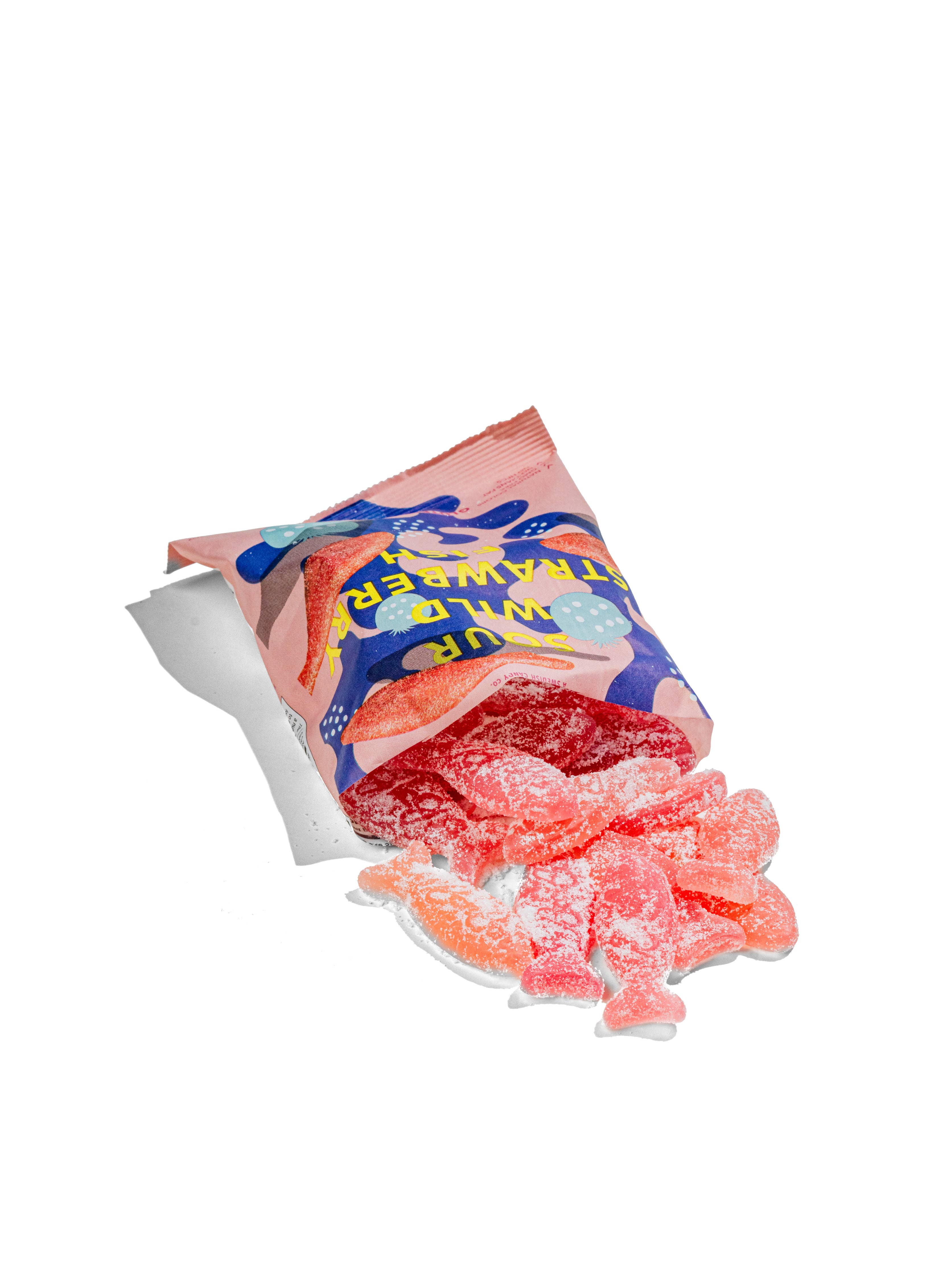 An open pink bag of BonBon - A Swedish Candy Co’s Sour Wild Strawberry Fish (5.2oz/150g) spills red, sugar-coated, vegan gummies shaped like fish onto a white surface—each bursting with wild strawberry flavor.