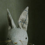 A close-up of the Skippy Cotton Ruffle Bun Ragdoll, a cotton-filled art doll in a cotton dress, showcases its charming hand-stitched details—stitched eyes, long ears with blue thread lines, and thin whiskers—against a neutral background.