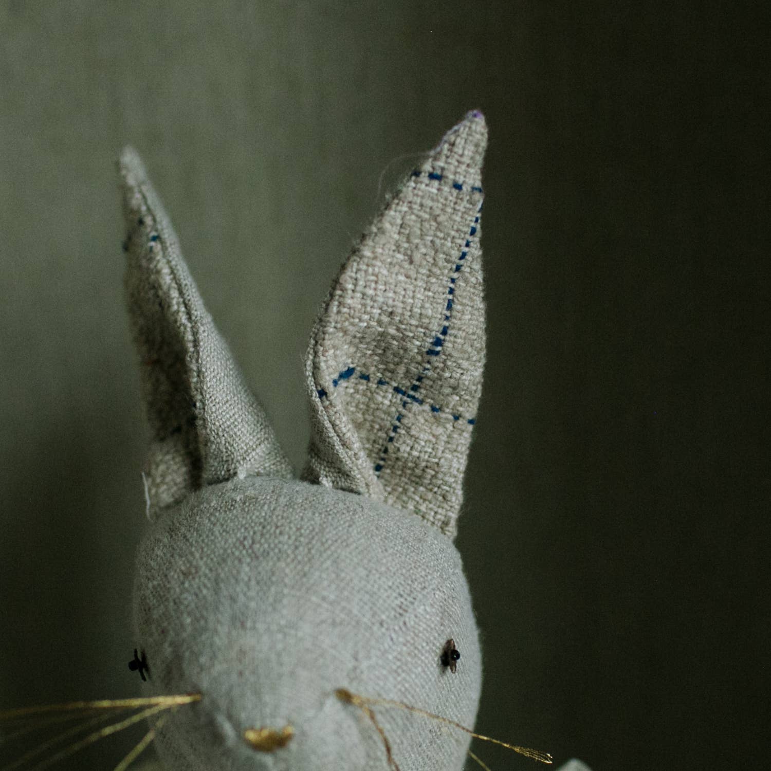 A close-up of the Skippy Cotton Ruffle Bun Ragdoll, a cotton-filled art doll in a cotton dress, showcases its charming hand-stitched details—stitched eyes, long ears with blue thread lines, and thin whiskers—against a neutral background.