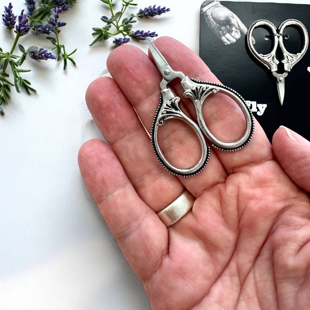 A hand holds ornate Firefly Notes Petite Thread Snips with decorative handles, beside lavender sprigs and an additional pair of these portable sewing scissors shown on a black card in the background.