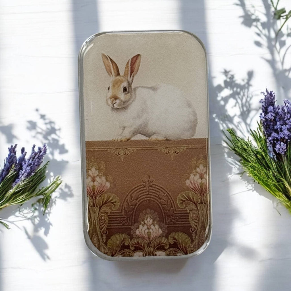 The Sweet White Bunny Notions Tin: Small by Firefly Notes features a vintage white rabbit on an ornate brown and gold background with purple flowers and green leaves, finished with glossy resin and photographed on a white surface.