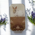 The Sweet White Bunny Notions Tin: Small by Firefly Notes features a vintage white rabbit on an ornate brown and gold background with purple flowers and green leaves, finished with glossy resin and photographed on a white surface.