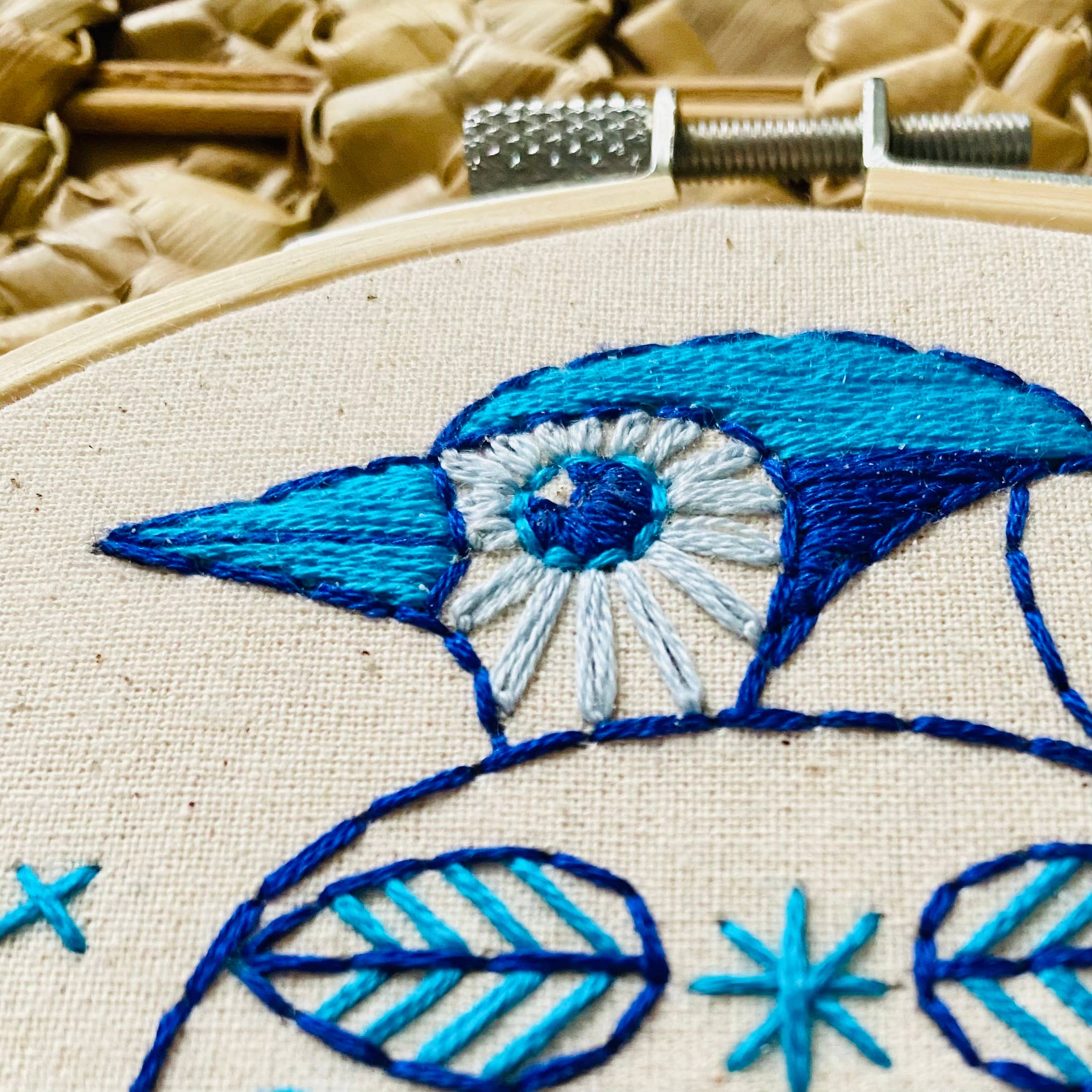 Close-up of the Blue Jay Complete Embroidery Kit by Hook, Line & Tinker Embroidery Kits, featuring a blue and white embroidered bird in a wooden hoop with textured woven background—ideal for beginners with detailed stitches and geometric accents.