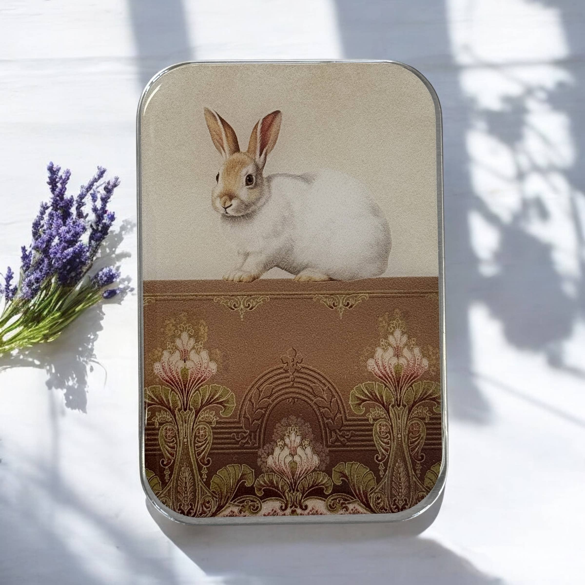 The Sweet White Bunny Notions Tin: Small by Firefly Notes features a unique illustration of a white rabbit on an ornate, vintage-style surface with lavender accents, set against a sunlit white background.