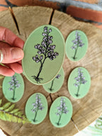 A hand holds an ilikesara | art + goods Iron-on patch | maidenhair fern, oval and green with a black and white floral design. Several similar patches are arranged on a round wooden surface in the background.