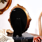 A Vintage Style Ornate Oval Table Mirror by Vagabond Vintage with a gold and black finish stands on a black book. Nearby are a lace cloth, pink artificial rose, and wooden chair; an antique gold mirror hangs on the wall behind.