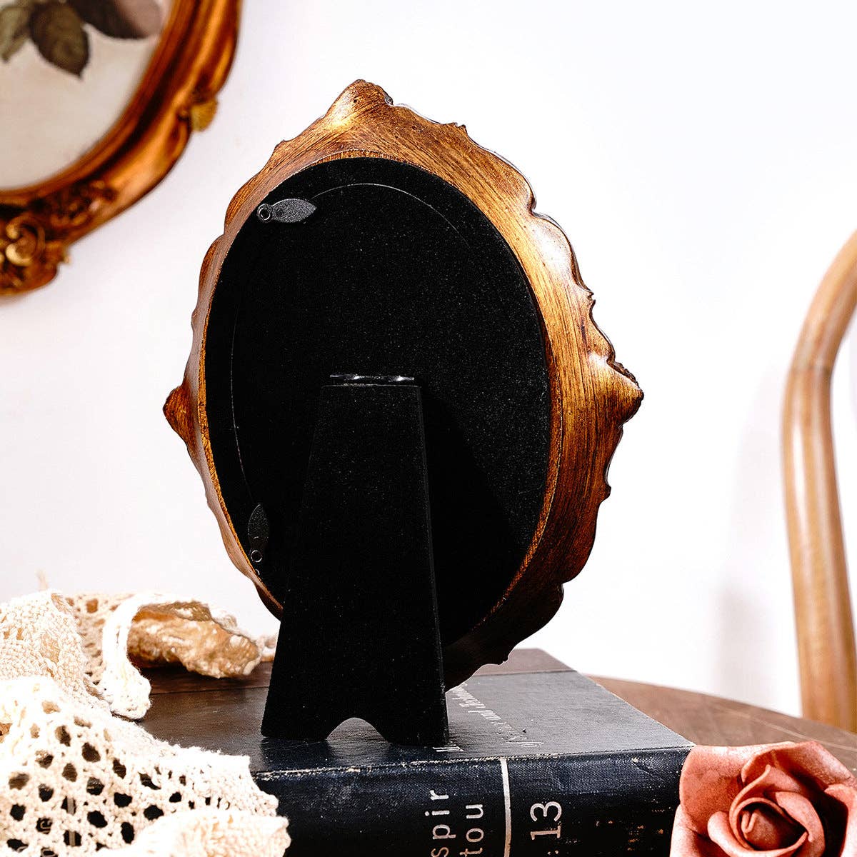 A Vintage Style Ornate Oval Table Mirror by Vagabond Vintage with a gold and black finish stands on a black book. Nearby are a lace cloth, pink artificial rose, and wooden chair; an antique gold mirror hangs on the wall behind.