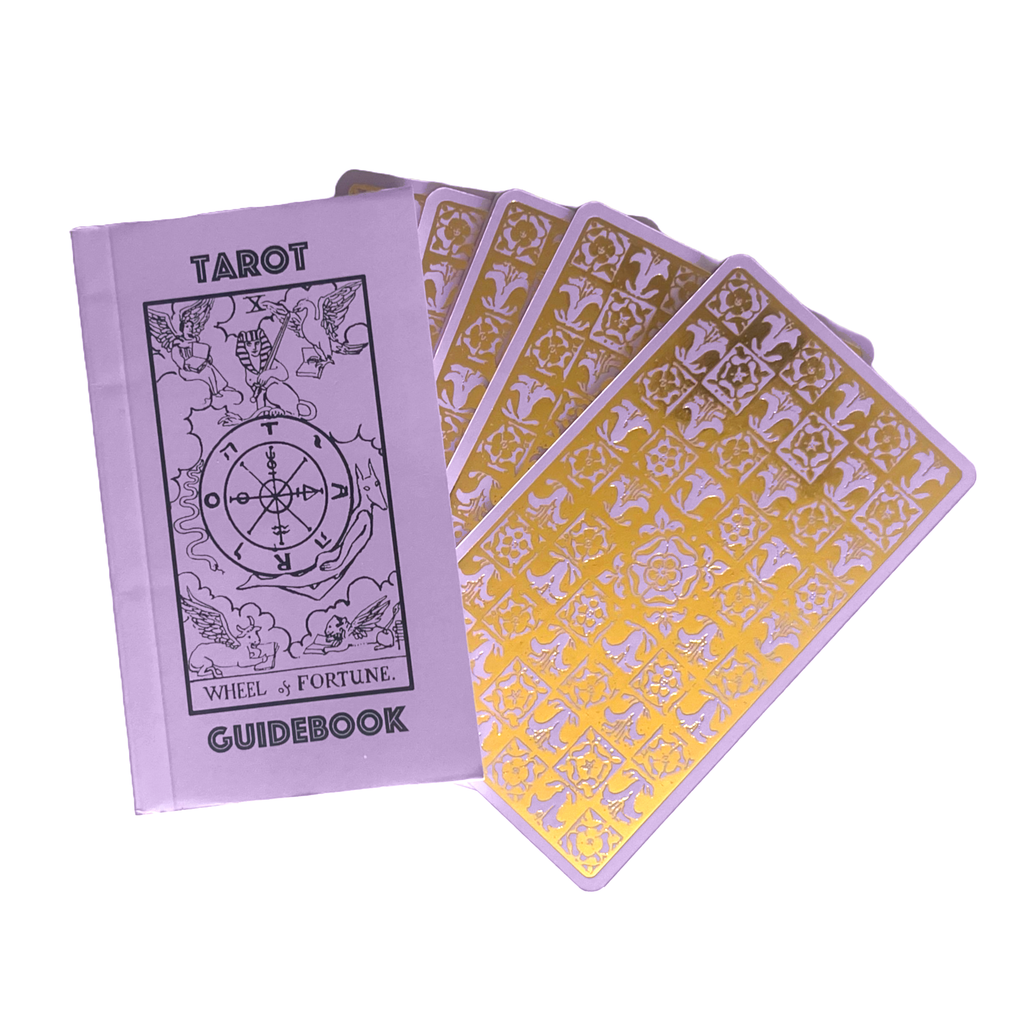 A Vieux Monde Lavender Gold Foil Tarot & Guide | Classic Tarot Design guidebook rests beside five tarot cards with ornate gold backs, fanned out on a white surface.