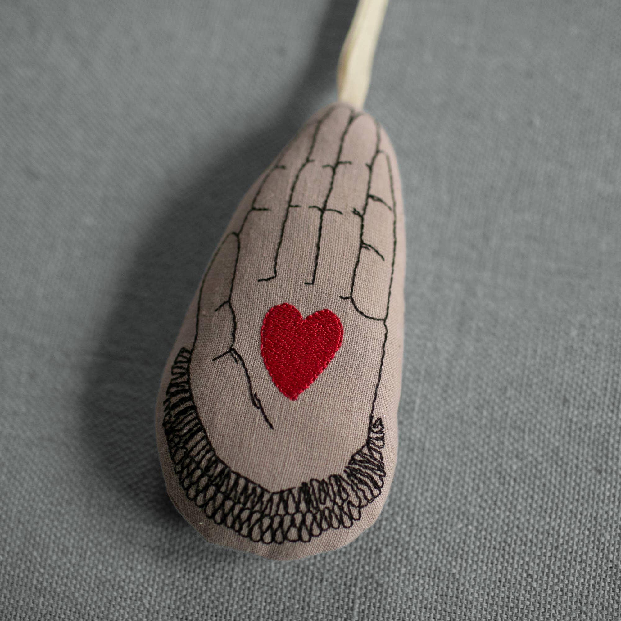 The Skippy Cotton Heart in Hand Ornament is an elongated teardrop-shaped organic cotton piece, embroidered with a hand and red heart, filled with lavender for scent, and set against textured gray fabric.