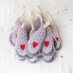 Skippy Cotton’s Heart in Hand ornaments, shaped like praying hands and filled with cotton and lavender, feature red hearts and black line details. They hang against a white background sprinkled with confetti.