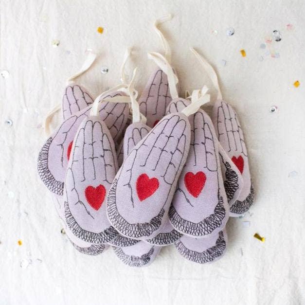 Skippy Cotton’s Heart in Hand ornaments, shaped like praying hands and filled with cotton and lavender, feature red hearts and black line details. They hang against a white background sprinkled with confetti.