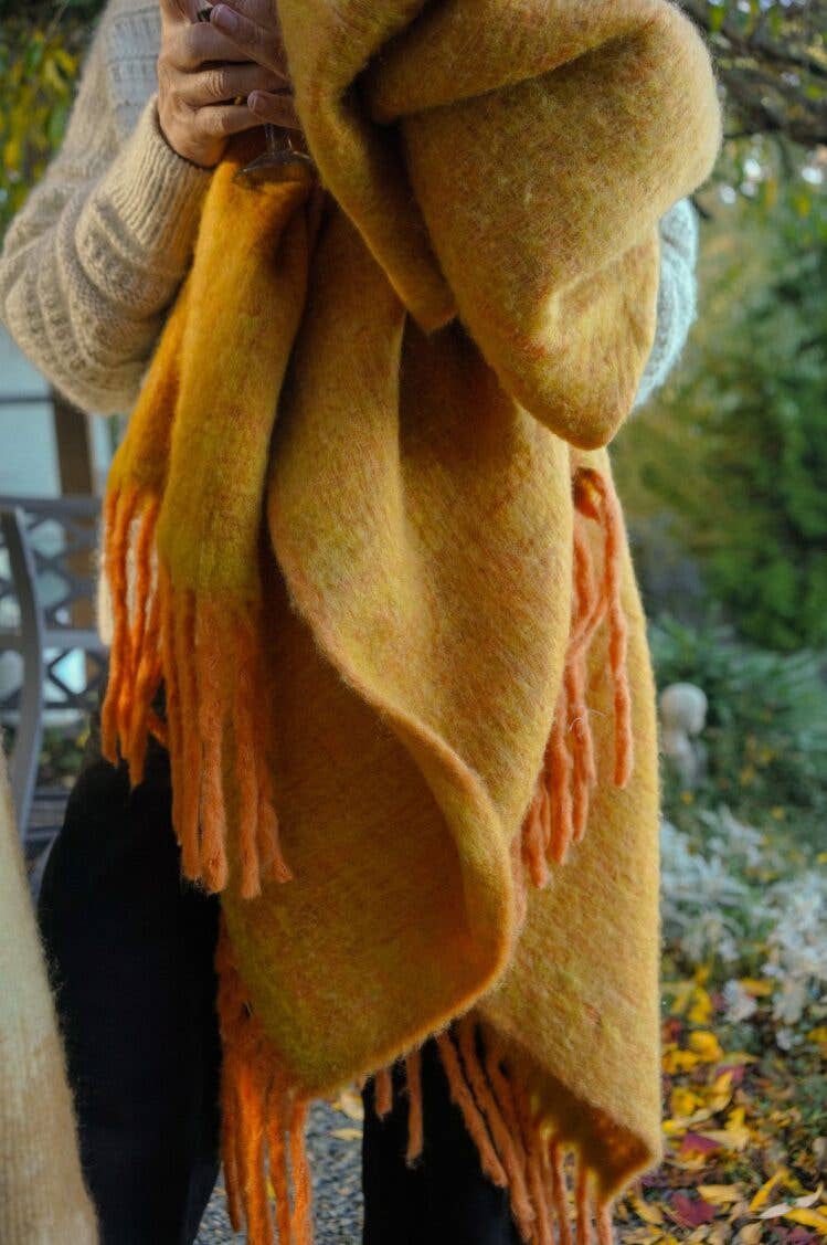 A person holds the Indaba Trading Oslow Wool Blend Throw in Turmeric—a large, fluffy, fringed blanket—outdoors among green foliage and autumn leaves, radiating cozy comfort.